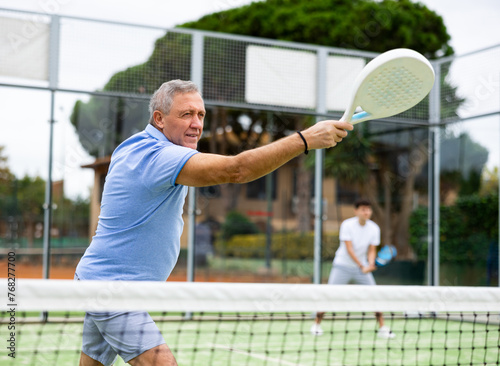 Focused aged man playing friendly paddleball match on outdoor summer court. Senior people sports concept.. © JackF