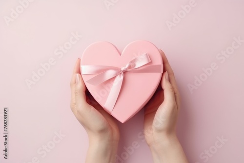 Hands holding a heart-shaped pink gift box with a satin ribbon on a light background. © Оксана Олейник