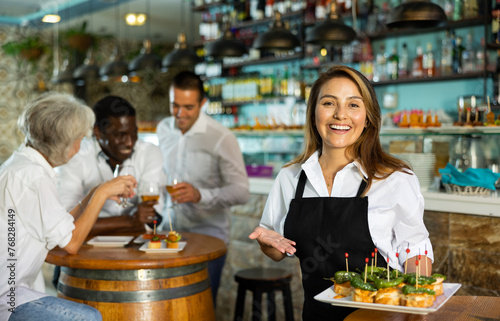 Happy Latin female waiter in uniform pointing hand on the plate with pinchos behind a table with diverse friends and bar counter