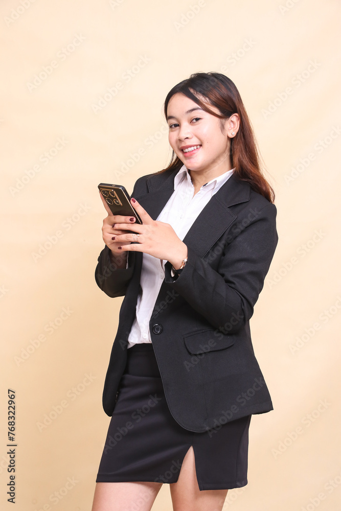 Beautiful formal Asian executive woman in suit standing cheerful smile and holding while operating online shopping gadget smartphone on yellow background for commercial, vacancy and business