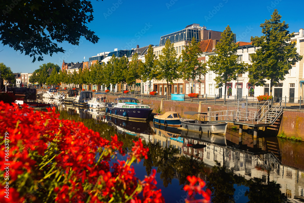 Mark River embankment in Breda, Netherlands. View of red flowers in bloom, buildings and boats along riverside.