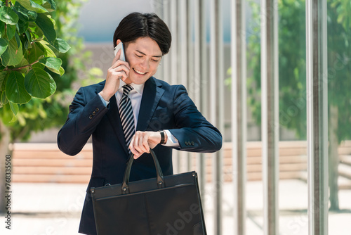スマホで通話しながら腕時計で時間を確認するスーツ姿の若いアジア人男性ビジネスマン
