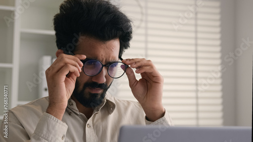 Tired overworked man Indian Arabian businessman working on laptop in glasses confused business employer taking off eyeglasses eyes problem blurry vision check eyesight eye laser correction health care © Yuliia