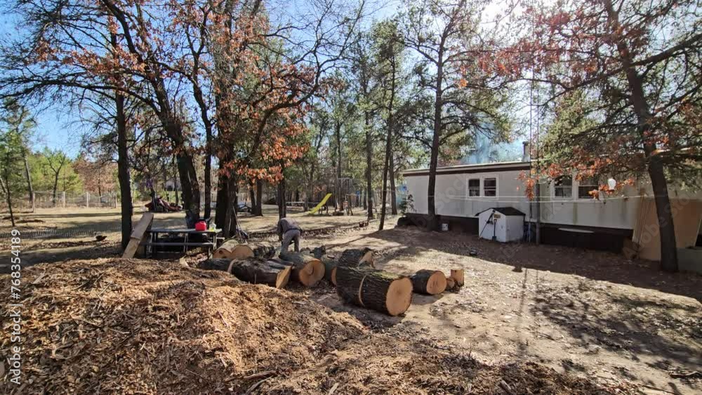 Wide angle shot of smoke rising from manufactured house chimney as wood being burnt in fireplace meanwhile skinny man in hood using chainsaw to process oak logs into firewood. Wide angle shot of smoke rising from manufactured house chimney as wood being burnt in fireplace meanwhile skinny man in hood using chainsaw to process oak logs into firewood.