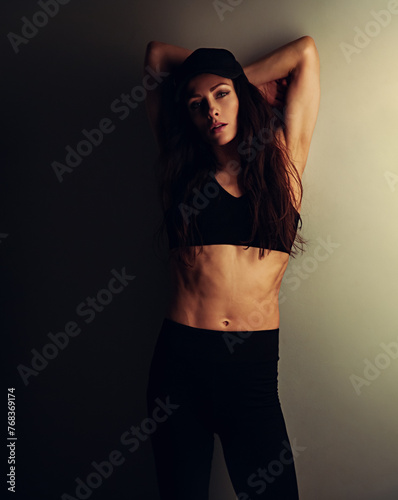 Sport sexy body beautiful slim woman with long hair posing in black sport bra, summer cap showing the shoulders, abs, arms, standing on studio wall background with empty copy space. Lifestyle vintage