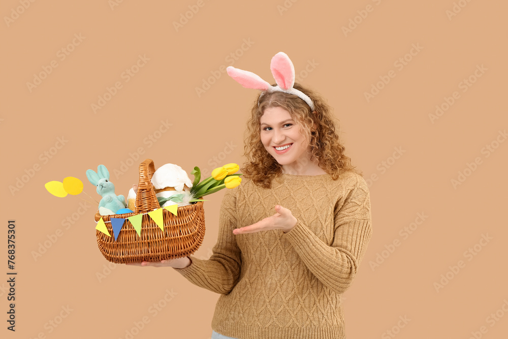Beautiful young happy woman in bunny ears pointing at Easter basket on beige background