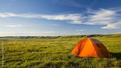 An orange tent pitched in the midst of expansive green hills under a blue sky with wispy clouds.