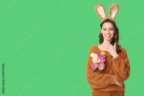 Happy young woman in Easter bunny ears headband with bouquet of tulips pointing at something on green background