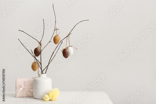 Branches with Easter eggs in ceramic vase, chick and gift near white wall