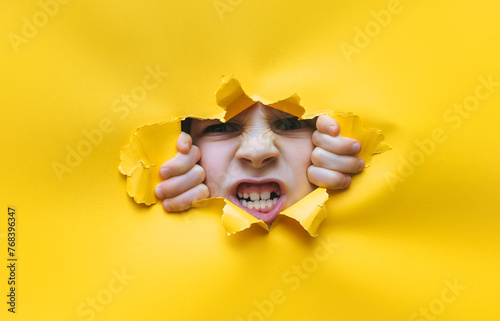 A little red-haired girl looks through a torn hole in yellow paper. The concept of anger, fear, fright and shock from what he saw. Unexpected aggression and stress. Copy space.