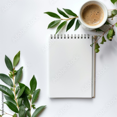 Blank white spiral notebook on table surrounded by coffee and green leaves, in flat lay photography style with minimalist aesthetic, taken from top view perspective on white background.
