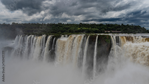 The incredible powerful waterfall - the Devil s Throat. Streams of foaming white water fall into the abyss. Green vegetation on the river bank. Clouds in the sky. Iguazu Falls. Argentina.