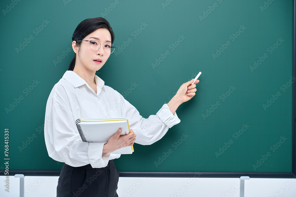 A beautiful Asian female teacher in white heating and glasses is holding a chalk in front of a green chalkboard and giving an Internet lecture with a confident expression and pose. A beautiful Asian female teacher in white heating and glasses is holding a chalk in front of a green chalkboard and giving an Internet lecture with a confident expression and pose.