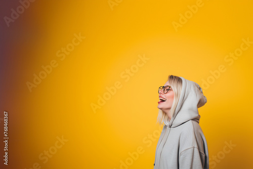 jeune femme blonde de profil, avec des lunettes de vue et portant un sweater à capuche gris, rigolant, personne jeune et heureuse sur un fond jaune moutarde dégradé avec espace négatif copy space