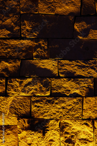 A grunge stone wall made of large textured bricks with bottom lighting, warm light, deep facture. Interior Design