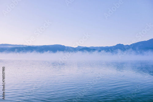 Picturesque scenery of the Lugu Lake in a thick white fog in sunrise. Silhouettes of mountains. Fall season. Nature, ecology, environmental conservation, eco tourism.