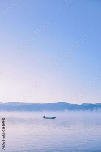 Picturesque scenery of the Lugu Lake in a thick white fog in sunrise. Silhouettes of mountains. A man floats in a boat with a paddle.