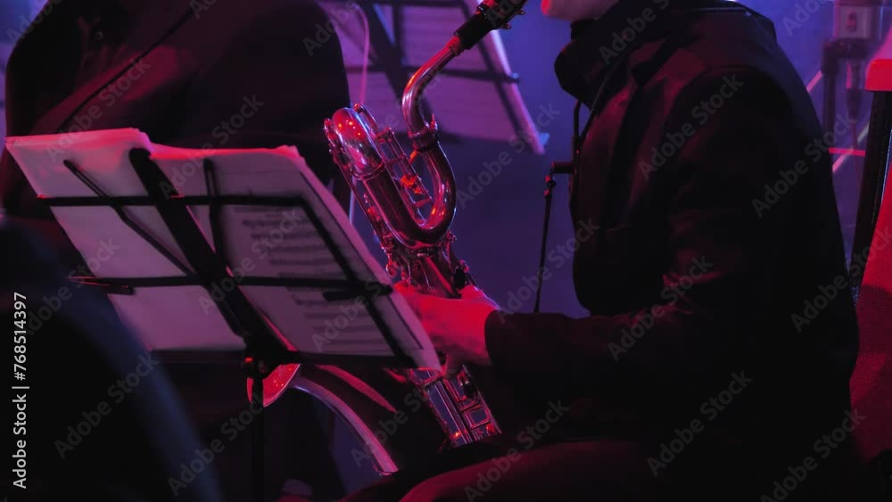 A musician in formal attire plays a baritone saxophone on a lit stage. A music stand with music sheets is visible. The atmosphere is dramatic and focused, emphasising the musician and his skill