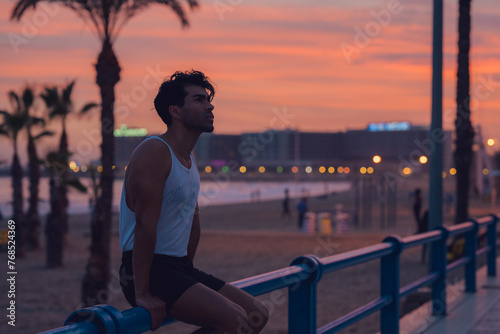 Pensive male athlete resting on a blue railing at beachside at sunset photo