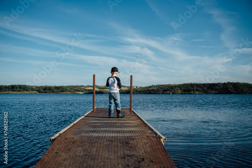 Wide rear view of small boy standing on dock looking out at lake.
