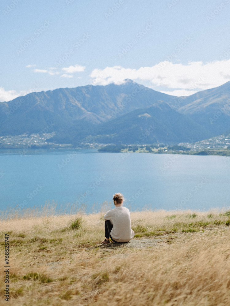 Woman sits on grassy overlook of blue lake and beautiful mountains Woman sits on grassy overlook of blue lake and beautiful mountains