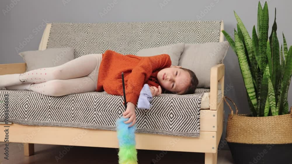 Tired little girl with brown hair being exhausted to help her mother with household chores lying on sofa with pp duster and rag yawning needs to have rest