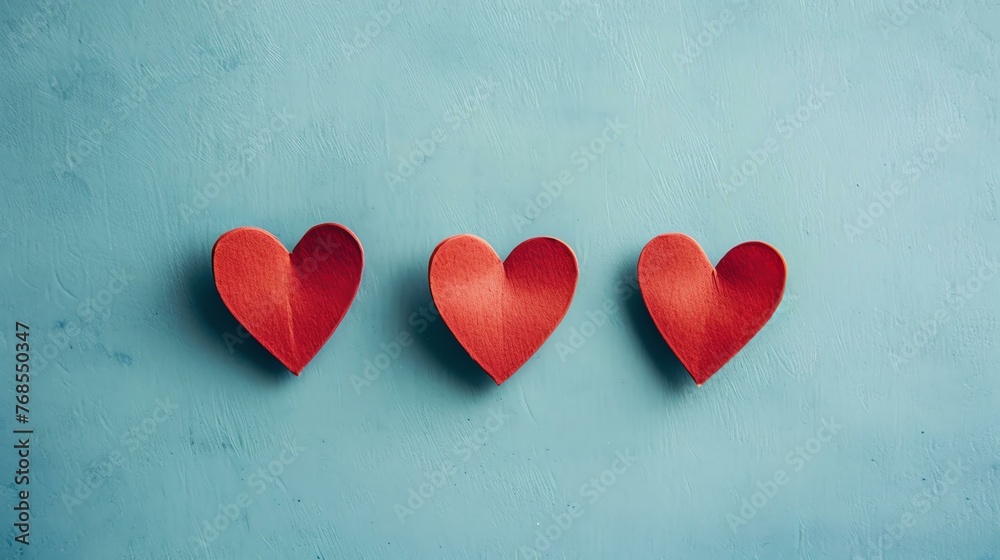 Three red paper hearts on a blue background. The hearts are arranged in a row. The background is a light blue color. The hearts are a deep red color.
