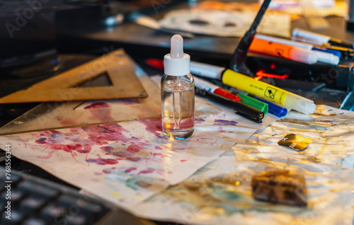 Workspace for art or crafts, with a small dropper-capped bottle containing a clear liquid in the center of the table. The surface is littered with various objects, including colored markers, a ruler, 