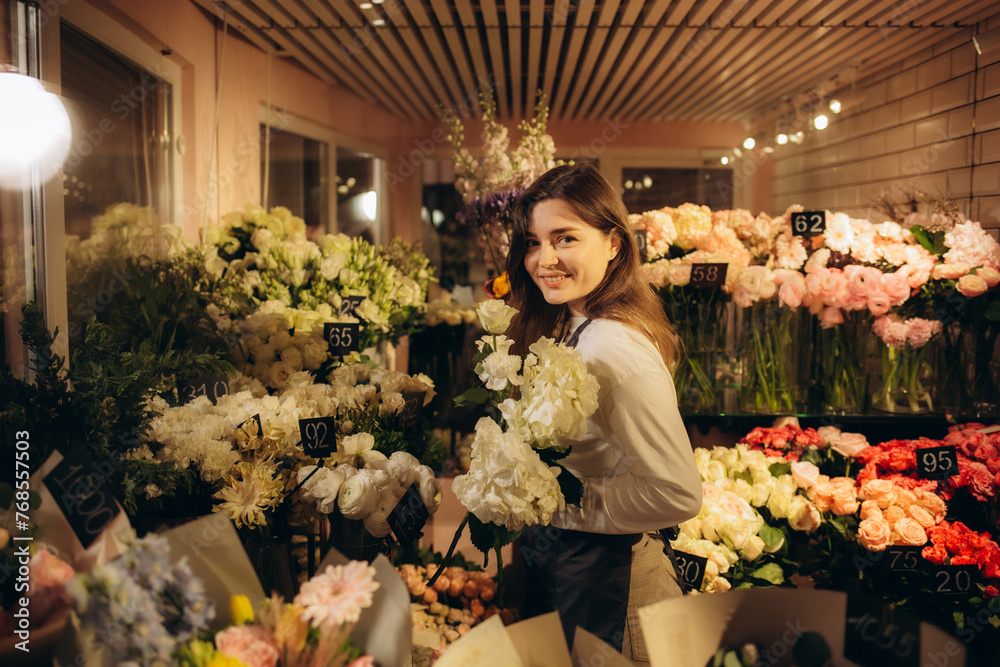 Cheerful young florist at work. Woman picking flowers for bouquet behind a glass in a flower storage room a refrigerator. Successful owner environment friendly concept copy space
