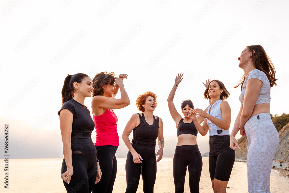 Obraz premium Group of adult athletic multiethnic women in sportswear have funny time at coast in background of sunset and sea. Concept of competition and female friendship