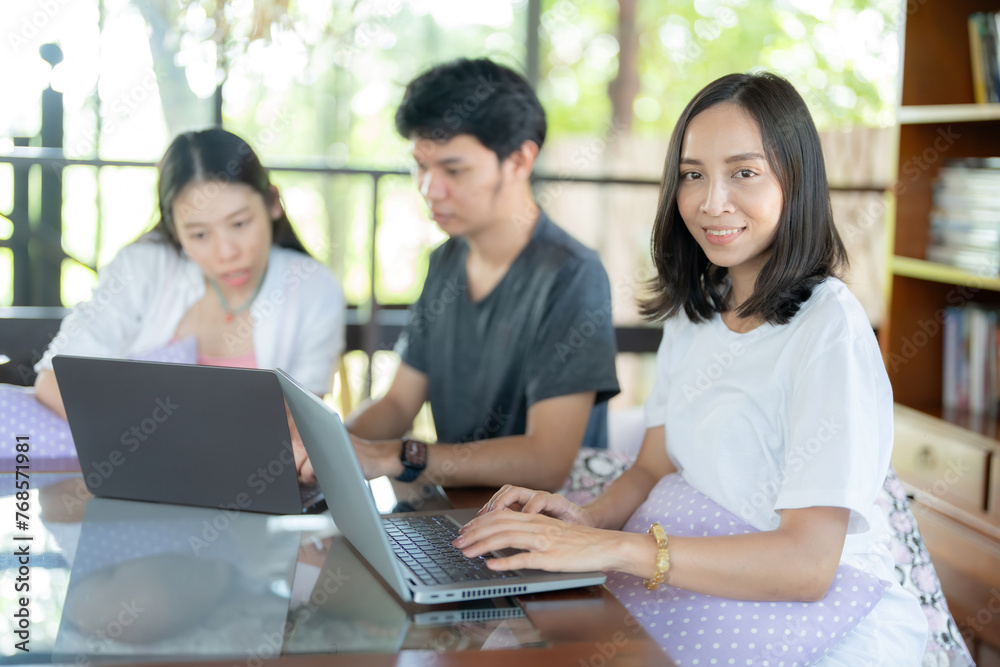 A female student with long Asian hair, beautiful and cute smile, uses a laptop to type work next to 2 friends working on a project. In the university café there is a library for research.