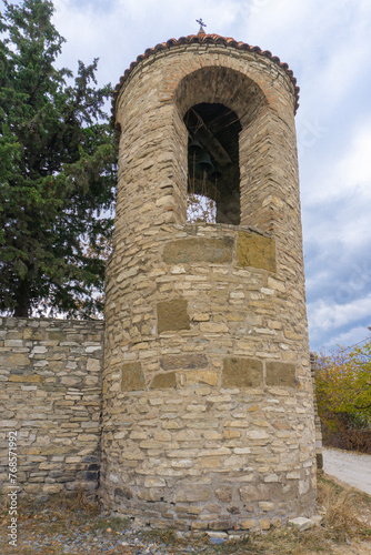 Bell tower and stone wall of Ertatsminda church. Bells are visible. Blue sky with clouds. Georgia