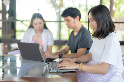 A long-haired Asian female student uses a laptop together with two friends to work on a project in the university cafe and the library where she uses her research to send to her professors.