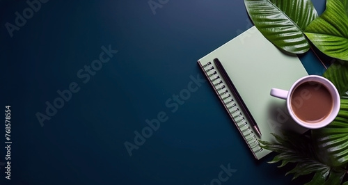 A closed paper notebook, black pen, green leaves of ornamental grass and a cup of coffee in a dark blue desk. View from the top. Image with copyspace for your text. Calm workspace concept. © bagotaj