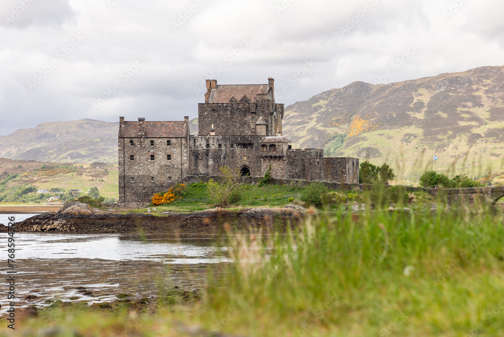 Eilean Donan Castle, a bastion of Scottish heritage, rises against a tapestry of mountainous terrain, with a foreground of lush grasses and a reflective tidal loch