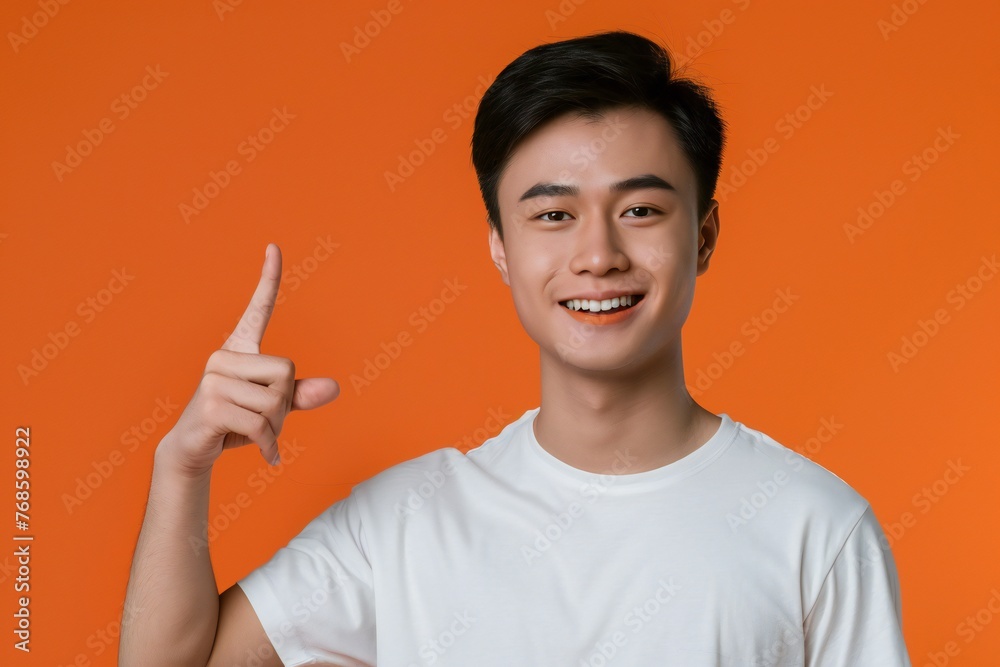 asian american male person with toothy smile on face pointing with index finger on empty space over orange background. Handsome young guy indicating place for advertising text in studio asian american male person with toothy smile on face pointing with index finger on empty space over orange background. Handsome young guy indicating place for advertising text in studio