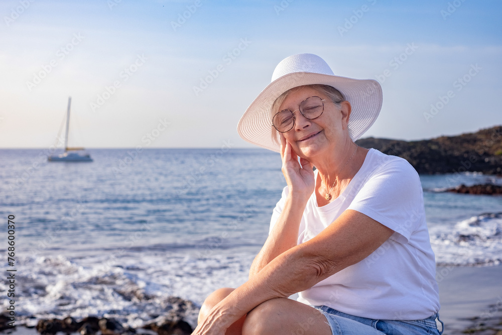 Smiling dreaming senior woman with white hat and closed eyes enjoying sea beach at sunrise, vacation or retirement concept Smiling dreaming senior woman with white hat and closed eyes enjoying sea beach at sunrise, vacation or retirement concept