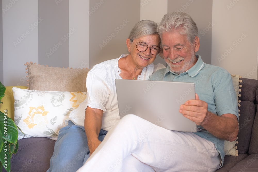 Cheerful senior retired couple sitting on sofa, using laptop technology, enjoying happy moments together in good mood and harmony Cheerful senior retired couple sitting on sofa, using laptop technology, enjoying happy moments together in good mood and harmony