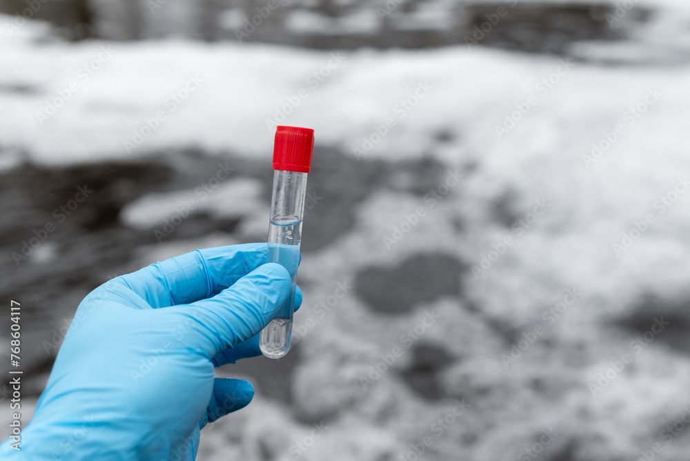 Fototapeta premium Test tube with red cap and water inside in male hand in blue medical gloves, close up. Foaming dirty water on the background, concept of water pollution