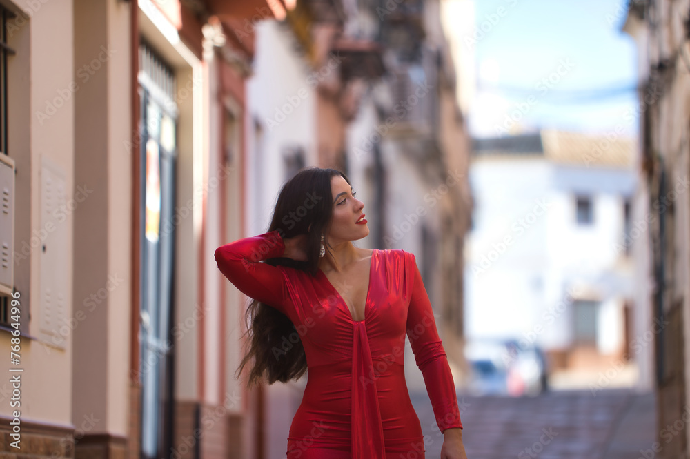 Young, beautiful brunette woman in an elegant red dress, walking while touching her hair, at dawn down a lonely street of a beautiful white Andalusian village. Concept beauty, fashion, trend, travel.