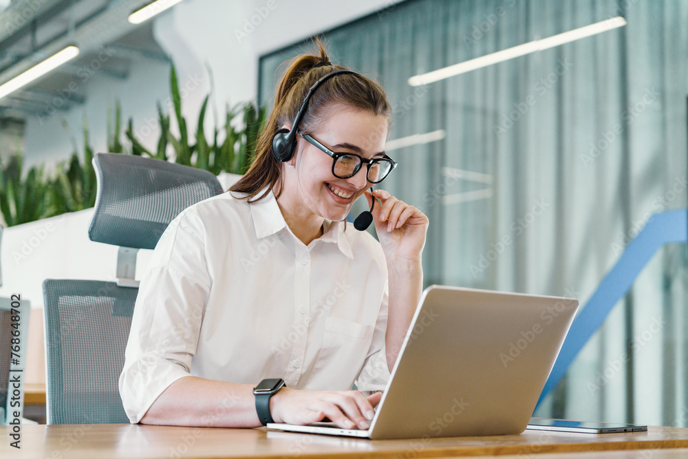 A genial customer support agent with a headset communicates cheerfully on a call while working on a laptop in an office with greenery. A genial customer support agent with a headset communicates cheerfully on a call while working on a laptop in an office with greenery.