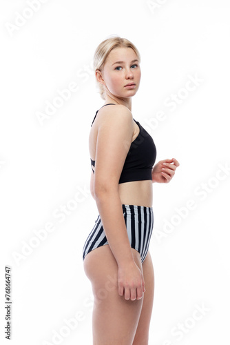 A young girl in a sports swimsuit. A cute blonde with freckles on her face in high striped panties and a black top. Isolated on a white background. Vertical.