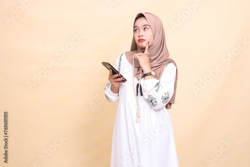 a young indonesia Muslim woman wearing a hijab thinks hard holding prayer beads and WhatsApp via smartphone gadget. for advertising, lifestyle, banners and Ramadan