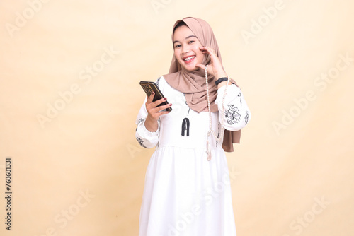 a young Asian Muslim girl wearing a hijab smiles in love holding prayer beads and chatting via smartphone gadget. for advertising, lifestyle, banners and Ramadan