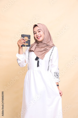 Beautiful mature Asian Muslim woman wearing a hijab with a cheerful expression looking at the camera carrying prayer beads tied to the Koran while holding a dress. for advertising and banners