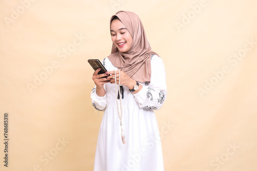 a young indonesia Muslim woman wearing a hijab smiles, laughs, holds prayer beads and chats via smartphone gadget. for advertising, lifestyle, banners and Ramadan