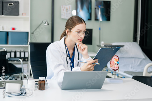 Wallpaper Mural Confident young Caucasian female doctor in white medical uniform sit at desk working on computer. Smiling use laptop write in medical journal Torontodigital.ca
