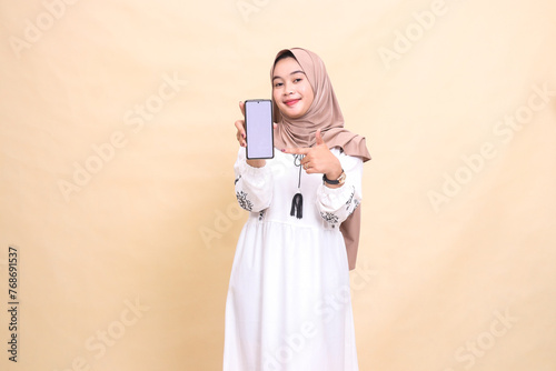 An adult Asian Muslim woman wearing a hijab smiles showing her cell phone (gadget) screen with a thumbs up gesture okay to create sales content. for advertising, technology, Eid and Ramadan