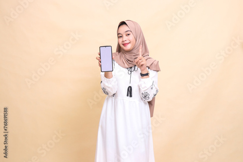 An adult Asian Muslim woman wearing a hijab smiles showing a cellphone screen (gadget) pointing up to create sales content. for advertising, technology, Eid and Ramadan