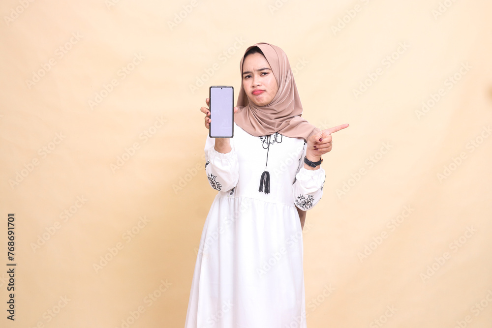 An indonesia Muslim girl wearing a hijab frowns showing a cell phone screen (gadget) pointing left to create sales content. for advertising, technology, Eid and Ramadan An indonesia Muslim girl wearing a hijab frowns showing a cell phone screen (gadget) pointing left to create sales content. for advertising, technology, Eid and Ramadan